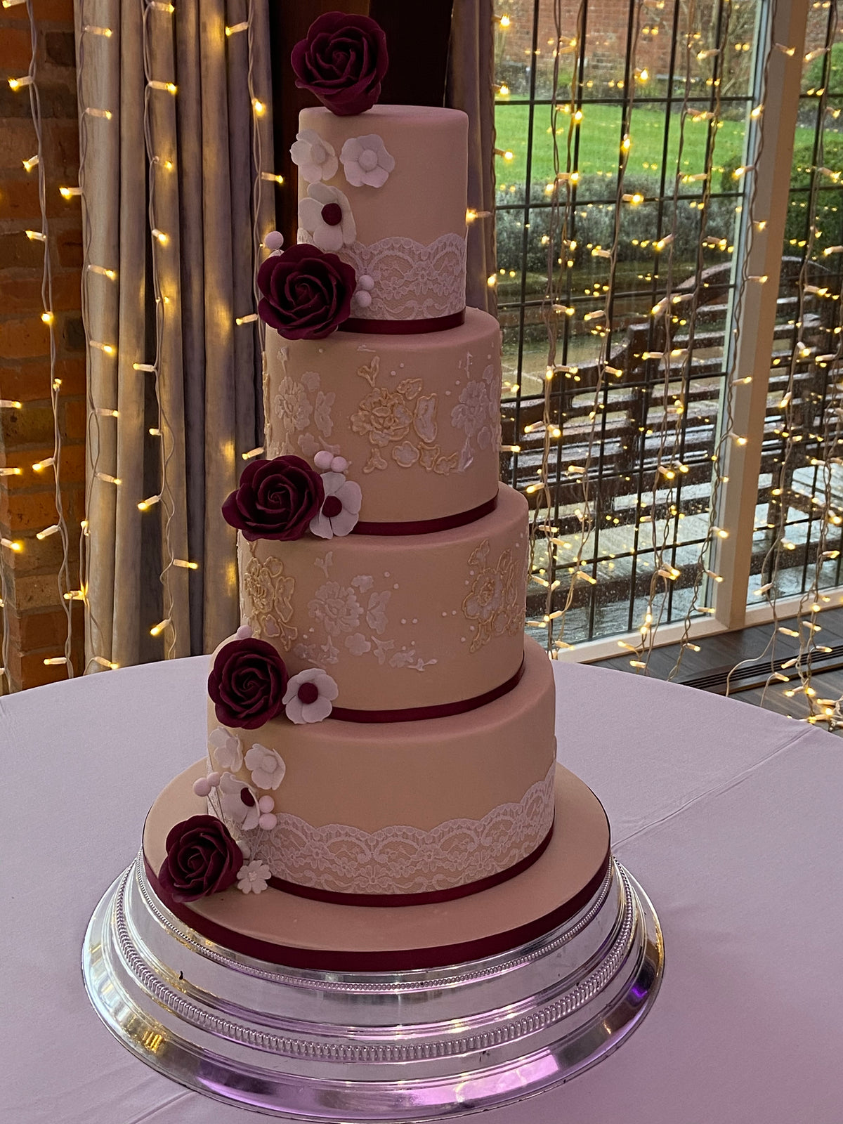 A four-tier peach wedding cake decorated with burgundy rose toppers, white floral accents and lace patterns, displayed on a silver cake stand in front of fairy lights