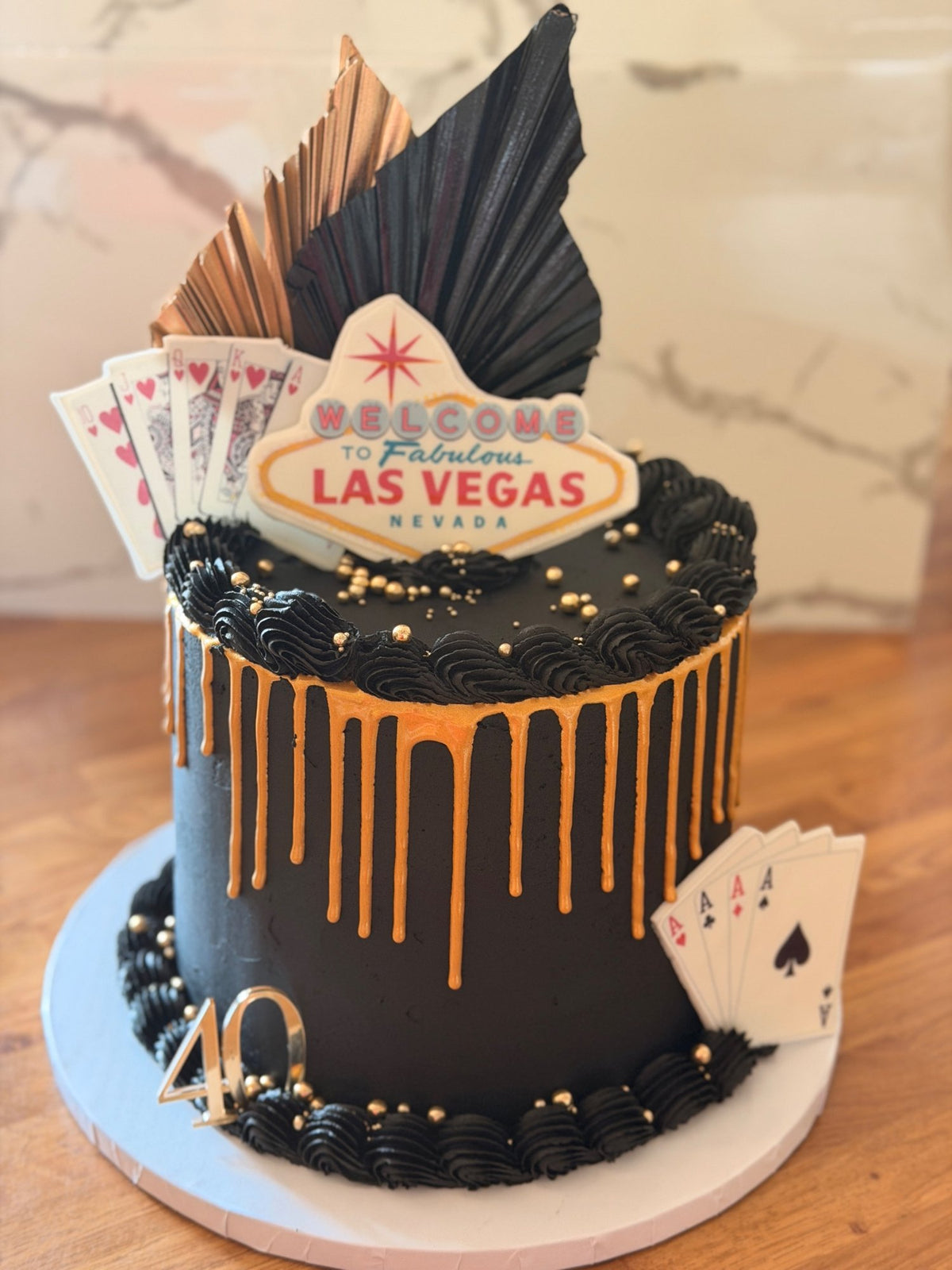 Black buttercream 40th birthday cake with a gold drip, a Las Vegas sign, playing cards, black and copper dried palm fans, gold sprinkles and a gold 40 topper