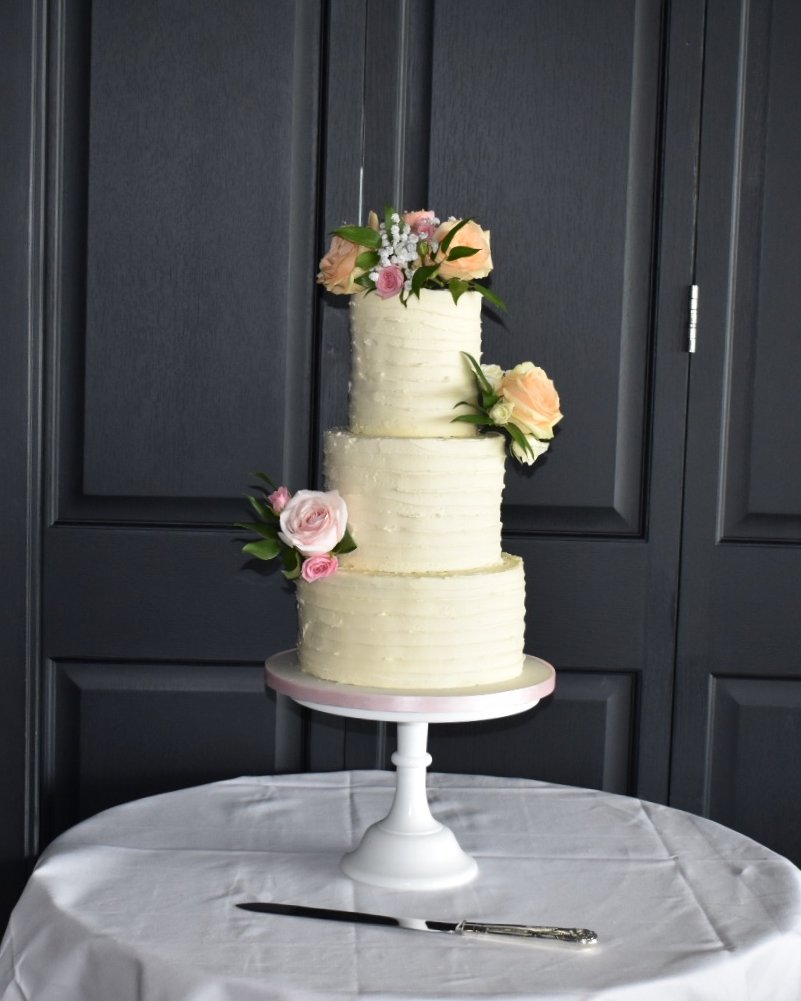 Three-tier buttercream wedding cake with a textured ridged finish, decorated with fresh peach and pink roses, on a white cake stand