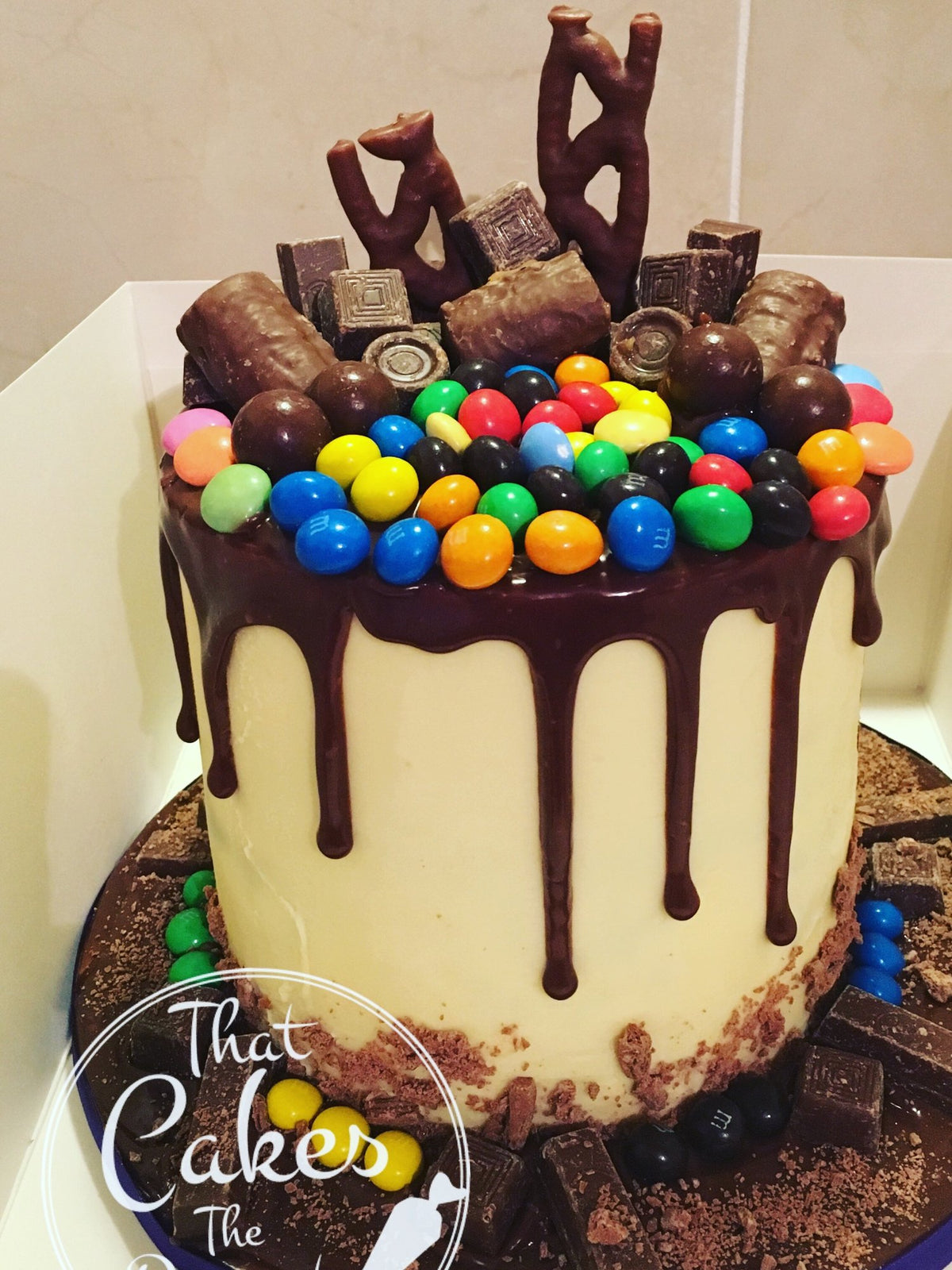 Tall white chocolate drip cake topped with M&Ms, chocolate bars and chocolate balls, with chocolate pieces and sweets around the base