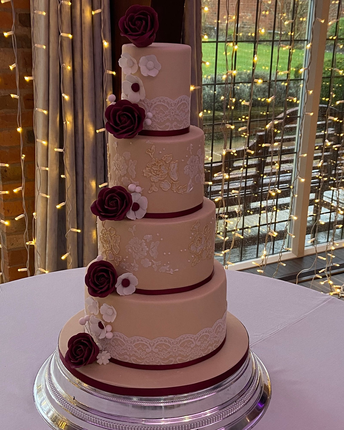 Four-tier blush pink wedding cake with white lace panels, hand-piped lace detail and a cascade of burgundy sugar roses and white blossoms, with burgundy ribbon on a silver cake stand in front of fairy lights