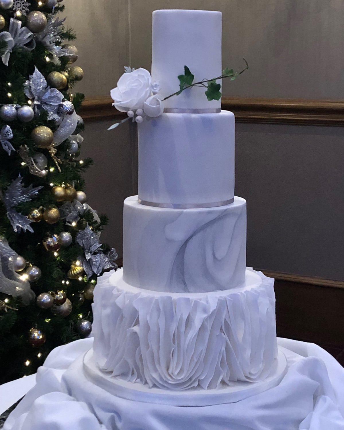 Four-tier wedding cake with marbled grey tiers, a white ruffled bottom tier, a white sugar rose and trailing ivy, beside a decorated Christmas tree