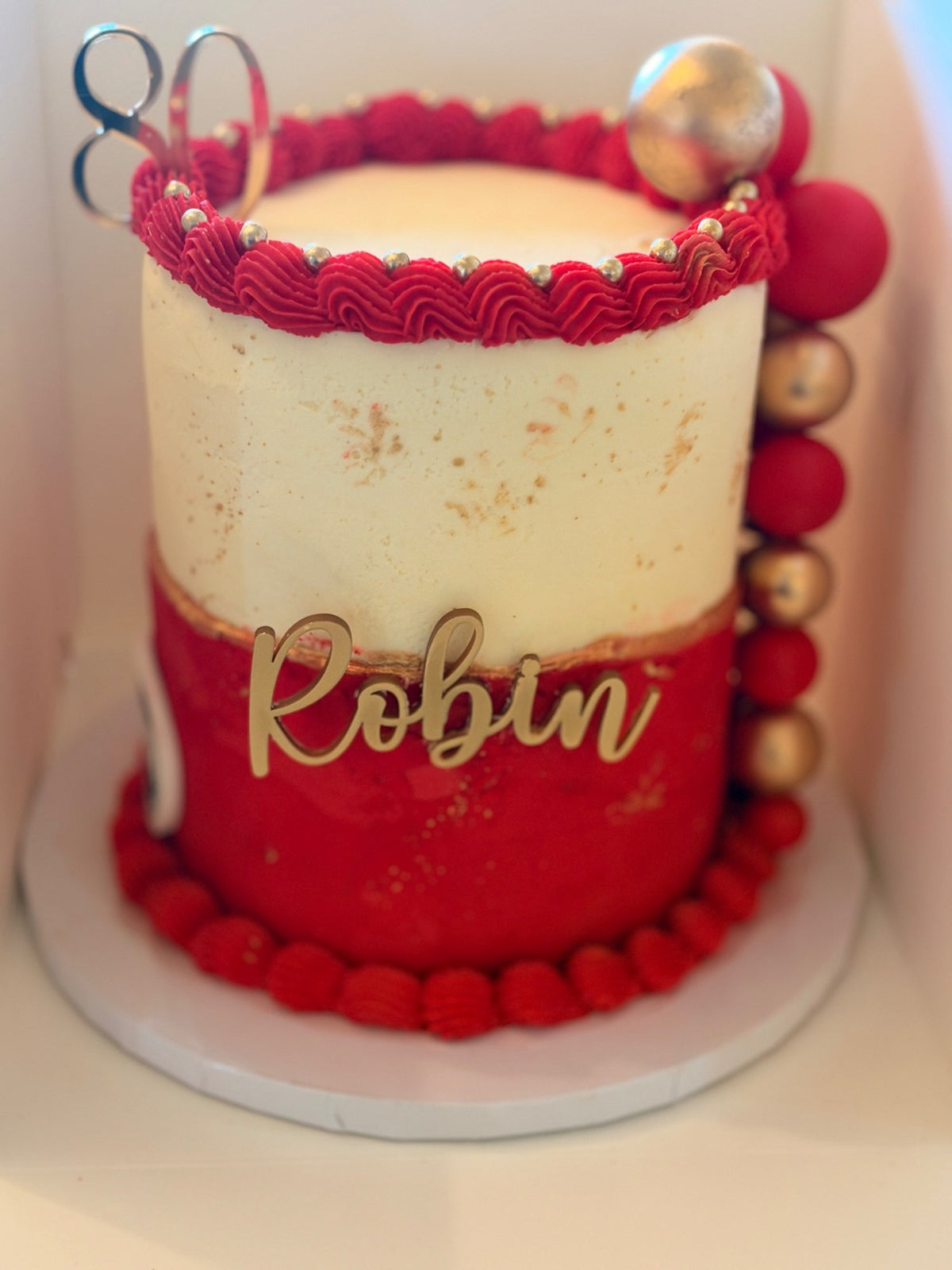 Tall 80th birthday cake with white buttercream above a red lower section, a red piped border, gold lettering for the name, a mirrored 80 topper and a column of red and gold spheres