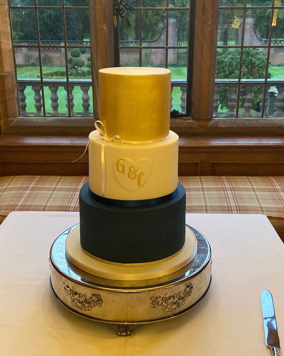 Three-tier wedding cake with a gold top tier, an ivory middle tier with a gold initials heart and a navy bottom tier, on a silver cake stand