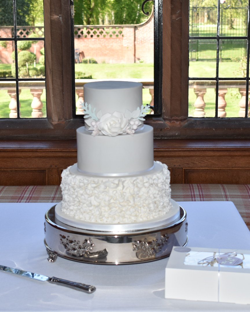 Three-tier wedding cake with smooth grey top tiers, a white ruffled bottom tier and a white sugar rose with soft green foliage, on a silver cake stand by a leaded window