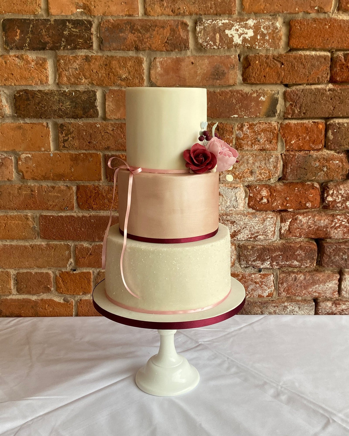 Three-tier wedding cake with an ivory top tier, a metallic rose gold middle tier and a sparkling ivory bottom tier, finished with a burgundy sugar rose, pink blossom and burgundy ribbon, on a white cake stand