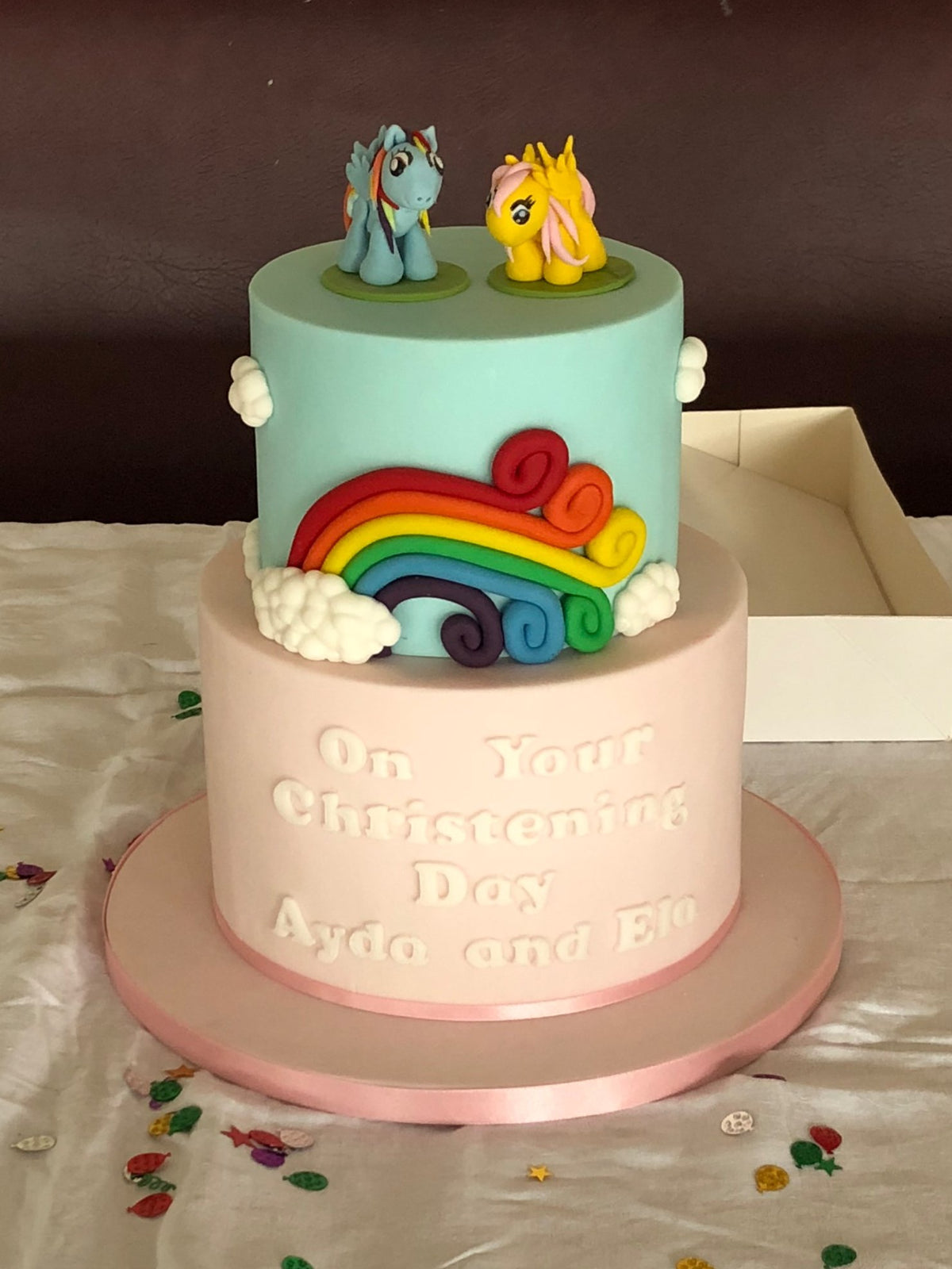 Two-tier christening cake with a mint blue top tier and a pale pink bottom tier, decorated with a fondant rainbow and clouds, two colourful pony figures on top and a personalised christening message