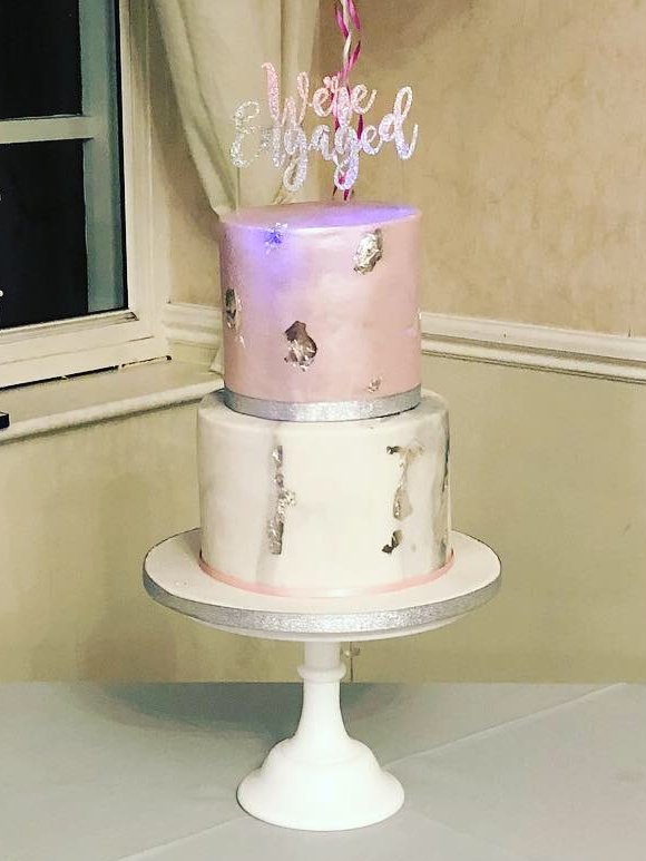 Two-tier engagement cake with a metallic pink top tier and a white marbled bottom tier, both finished with silver leaf, topped with a glittery We're Engaged sign on a white cake stand