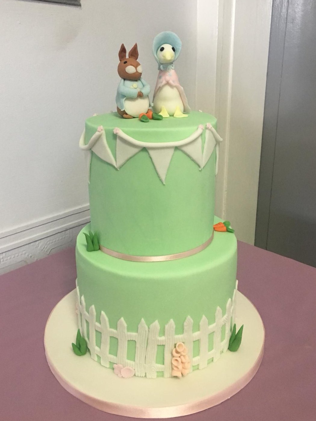 Two-tier pale green cake with Beatrix Potter style rabbit and duck figures on top, bunting, a white picket fence, and fondant carrots and grass, on a cream board with pink ribbon