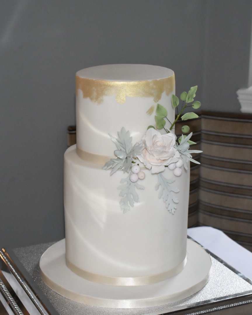 Two-tier white wedding cake with gold leaf on the top tier, finished with a sugar rose, dusty miller foliage, berries and eucalyptus